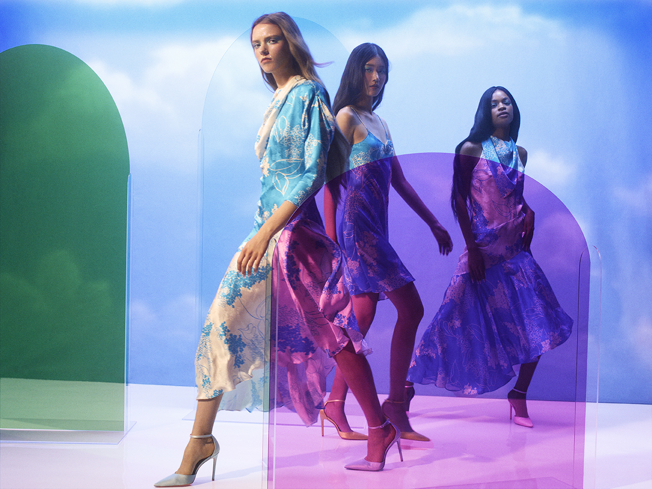 Three fashion models in floral dresses and high heels stand in a photo studio with colorful transparent partitions against a cloud background.