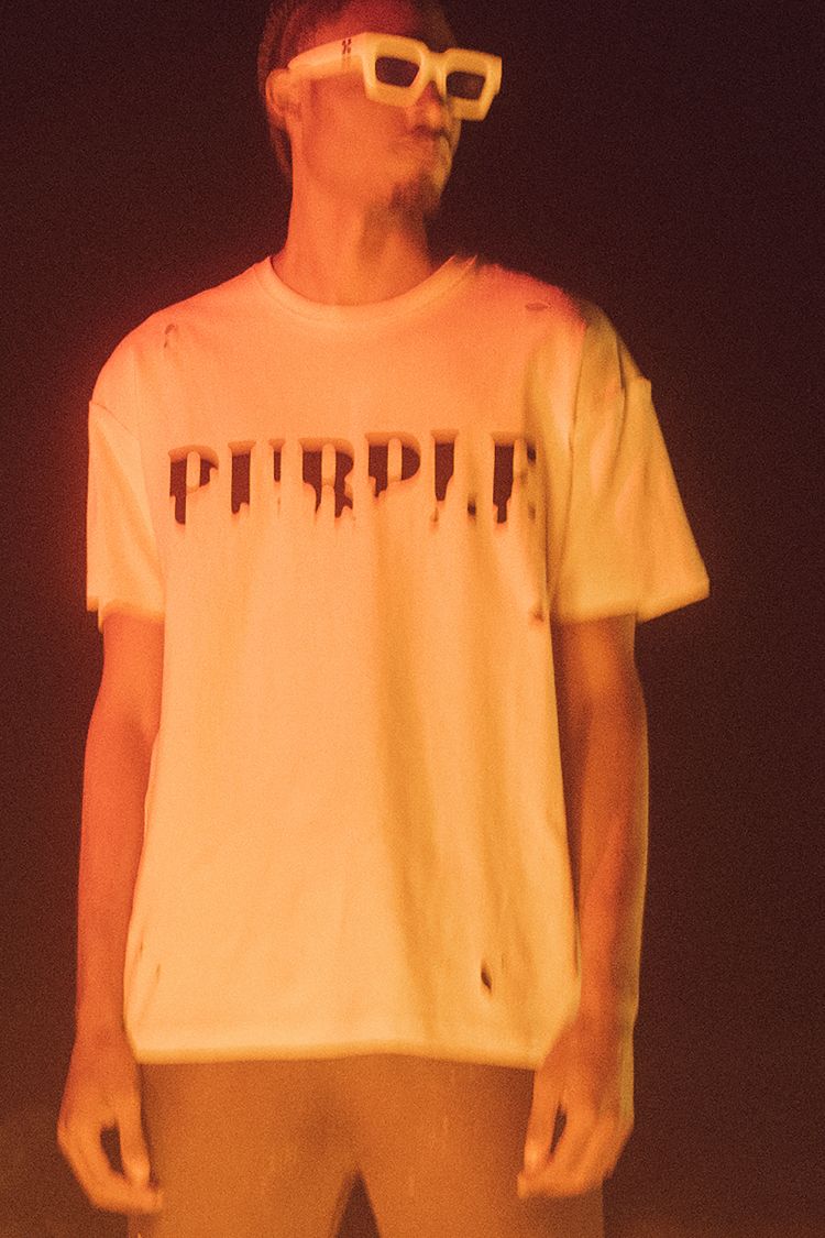 Man in white t-shirt and sunglasses posing against a dark background with warm lighting, showcasing fashion and personal style.