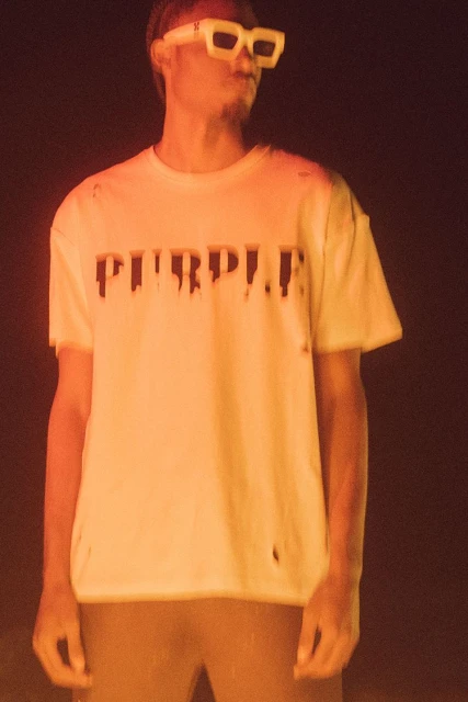 Man in white t-shirt and sunglasses posing against a dark background with warm lighting, showcasing fashion and personal style.