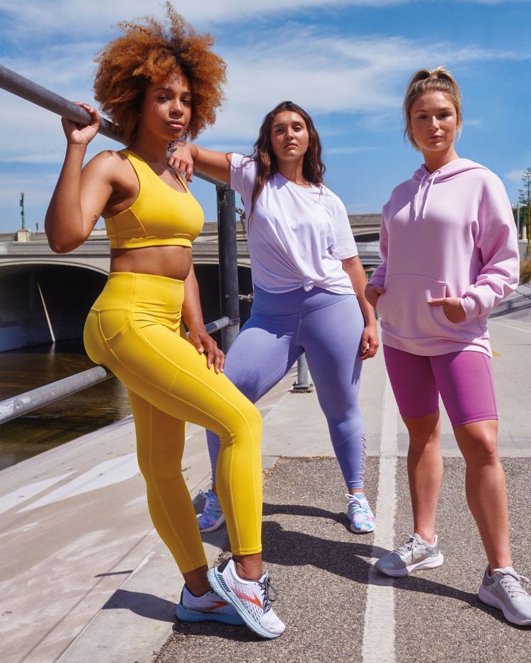 Three women in activewear pose outdoors by a river, showcasing yellow, lavender, and pink athletic outfits against a blue sky backdrop.