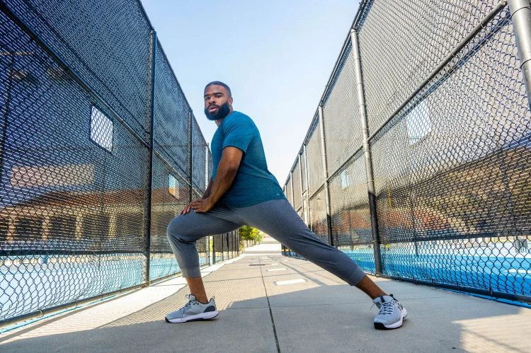 Athletic man in blue shirt and grey sweatpants stretches outdoors between chain-link fences, preparing for a fitness workout in daylight.