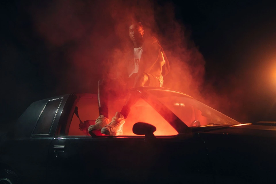 Young man sits on a classic car hood in a smoky, low-lit scene, bathed in red and orange light, creating a dramatic automotive portrait.