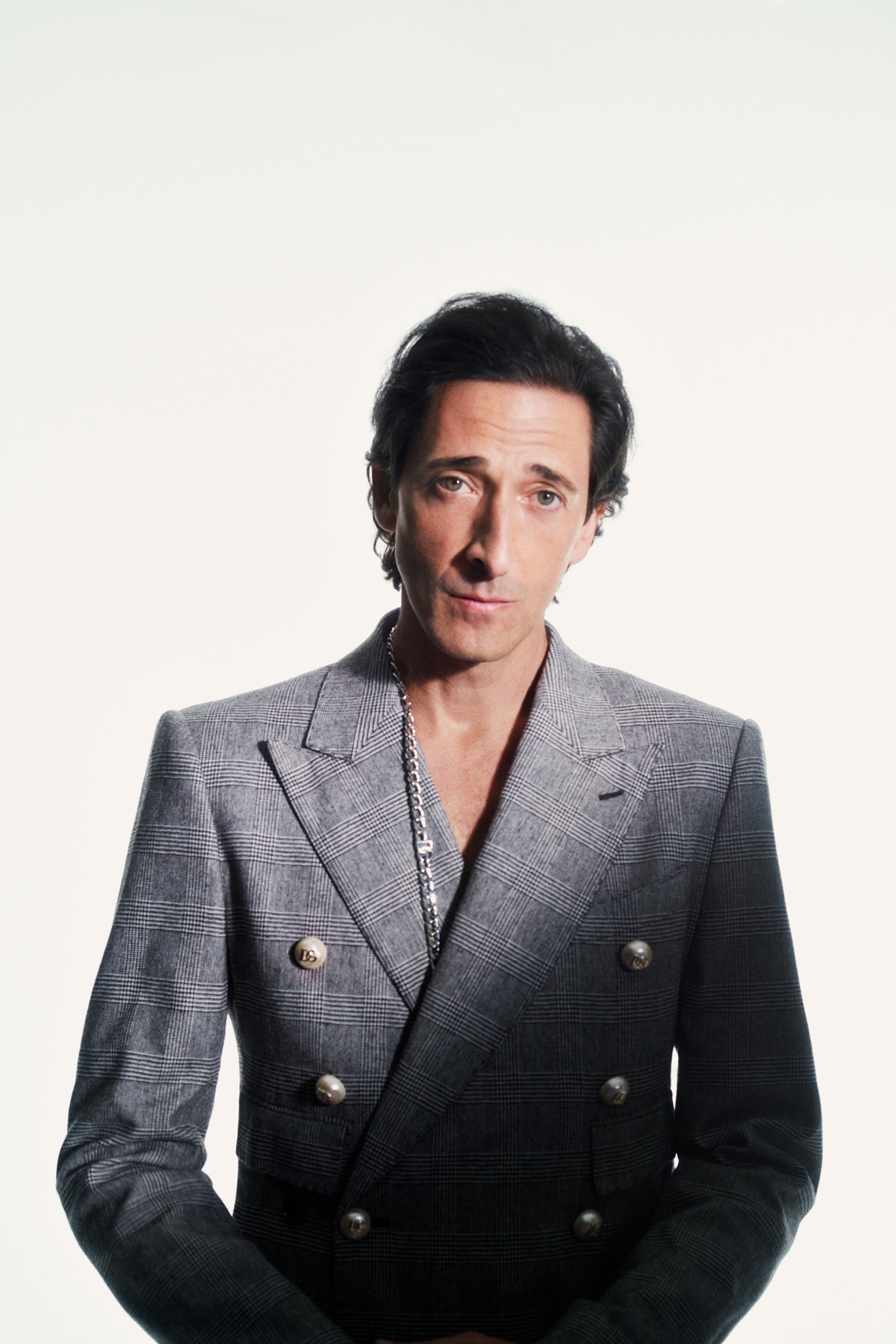 Portrait of a stylish adult male wearing a gray double-breasted blazer with metal buttons and chain, in front of white backdrop.