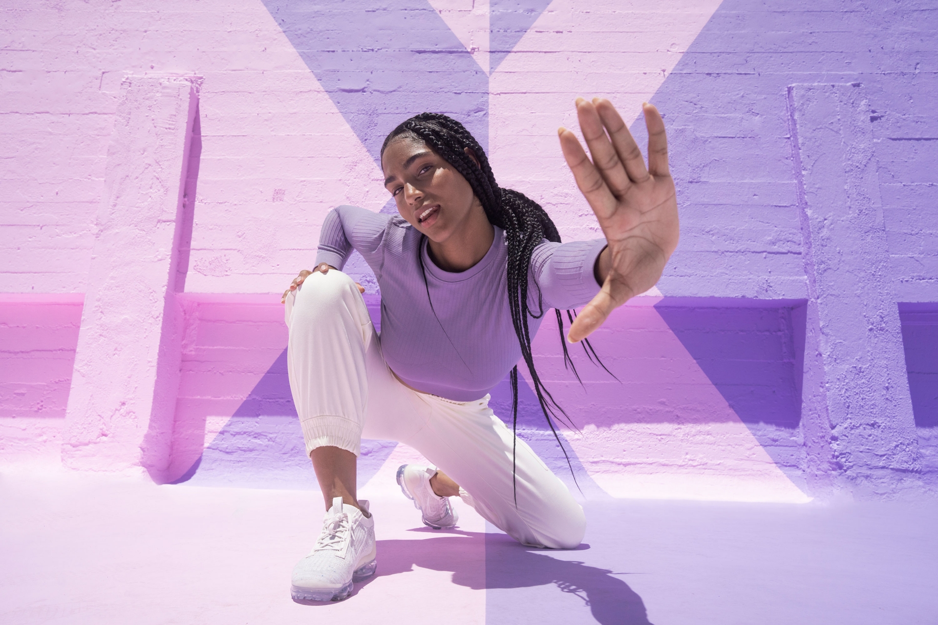 Young Black woman in athletic wear poses on a purple and pink surface in a studio with sunlit geometric wall patterns.