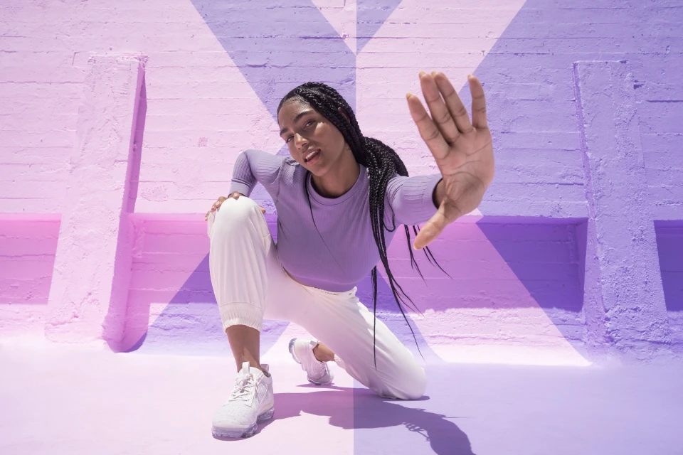 Young Black woman in athletic wear poses on a purple and pink surface in a studio with sunlit geometric wall patterns.