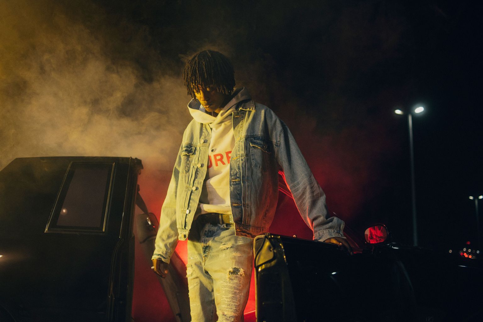 Young man with dreadlocks wearing a denim jacket and white hoodie standing next to a car at night with red smoke