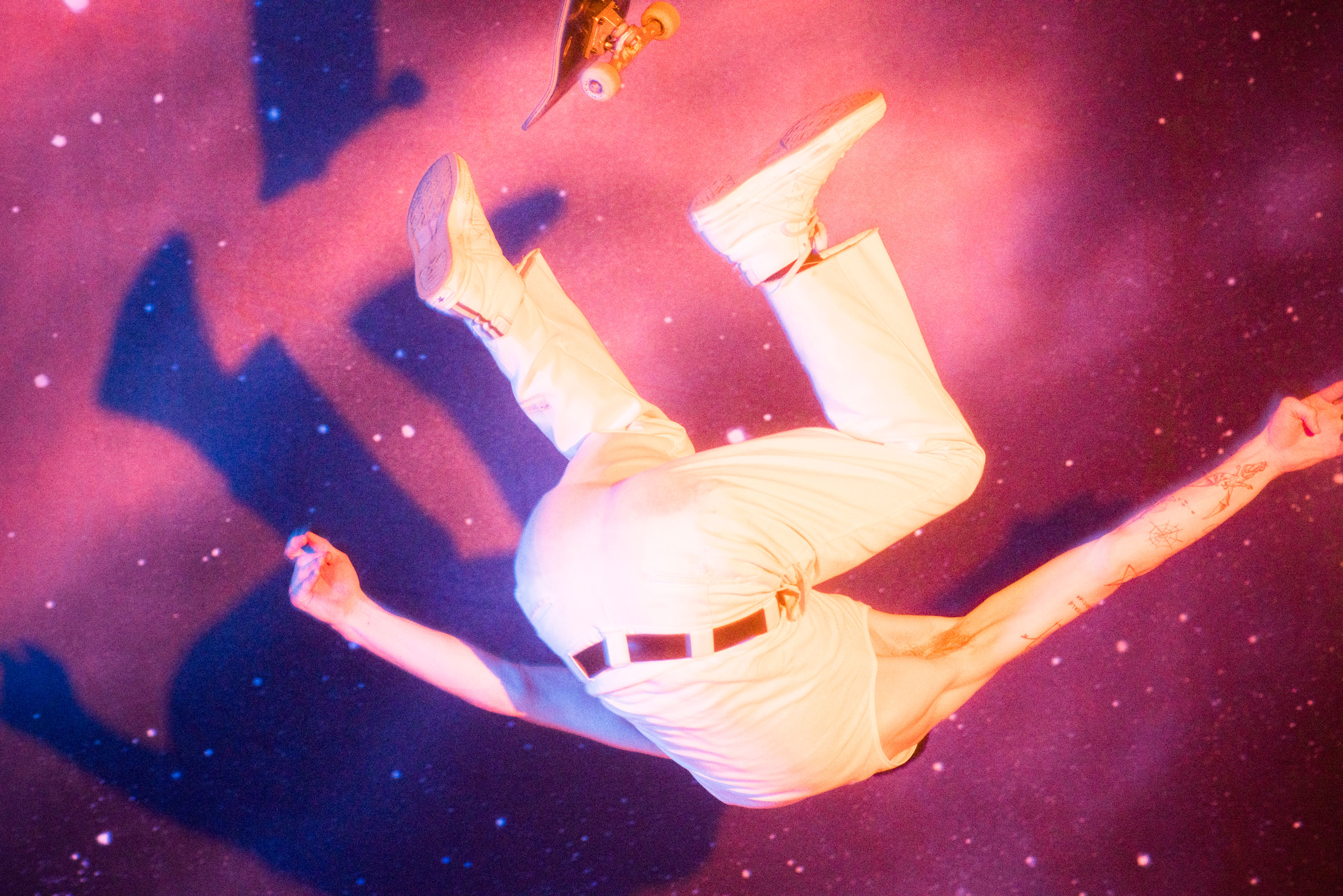 Man in white outfit appears to be falling in a studio with cosmic background and a skateboard, captured with vibrant pink and blue lighting.