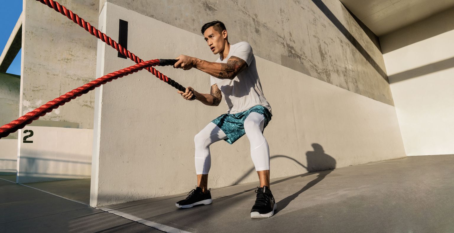 Athletic man in activewear works out with red and black battle ropes outdoors against a grey concrete wall on a sunny day.