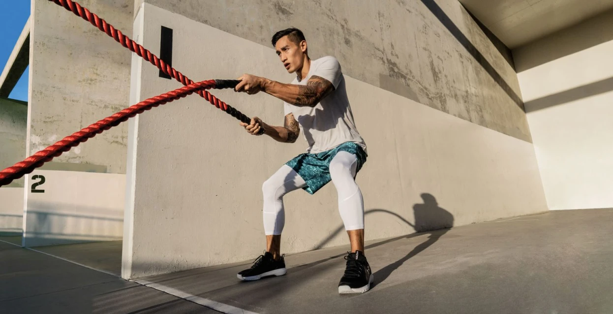 Athletic man in activewear works out with red and black battle ropes outdoors against a grey concrete wall on a sunny day.