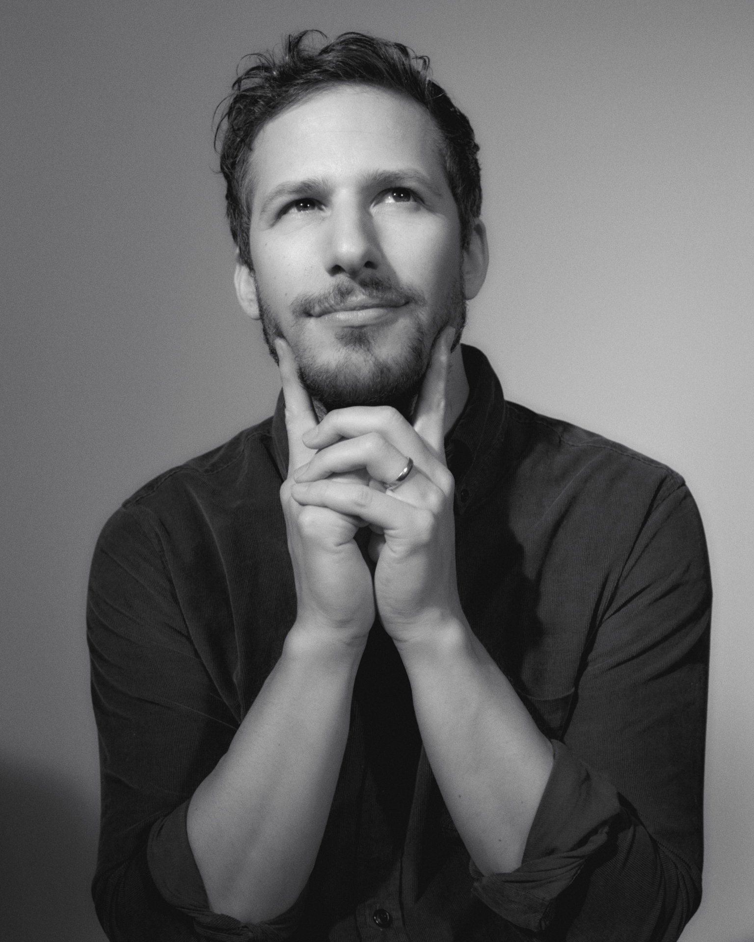Black and white studio portrait of an adult male with beard, looking up with hands touching face, wearing a button-down shirt.