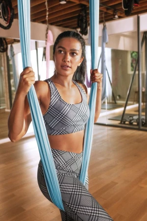 Adult female in a plaid workout set poses inside a fitness studio while holding onto a light blue aerial yoga hammock.