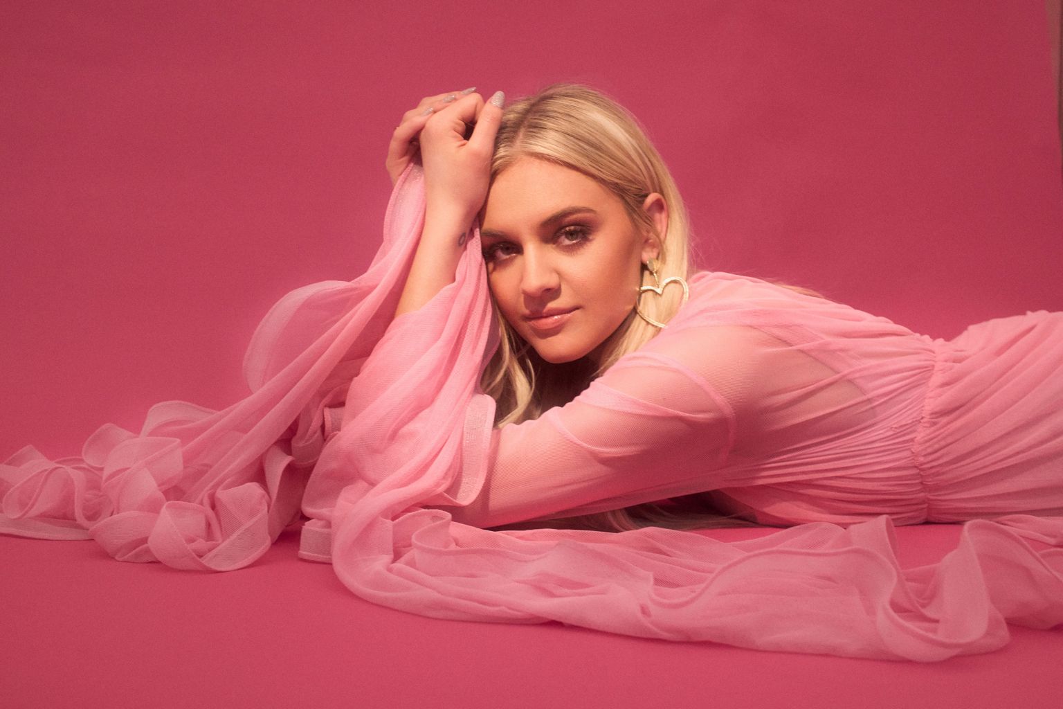 Blonde woman in pink dress lies against a pink backdrop. She wears heart earrings and has her hands above her head.