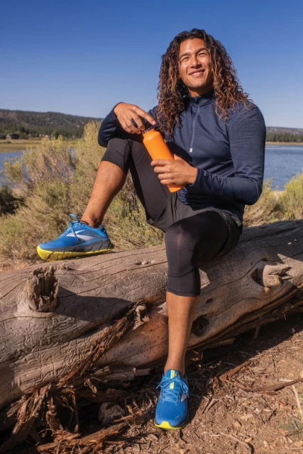 Adult male with long curly hair sits on fallen log and drinks water from orange bottle near lake on sunny day in athletic wear.