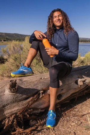 Adult male with long curly hair sits on fallen log and drinks water from orange bottle near lake on sunny day in athletic wear.