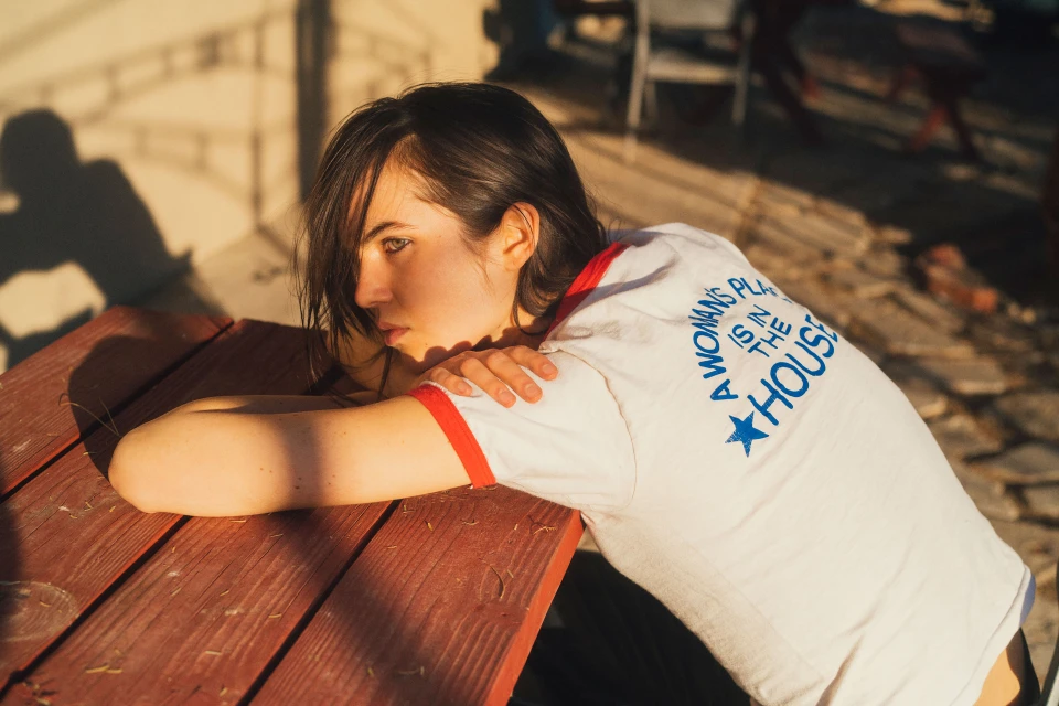Young woman rests on a red picnic table outdoors, wearing a slogan t-shirt, bathed in warm sunlight casting shadows.