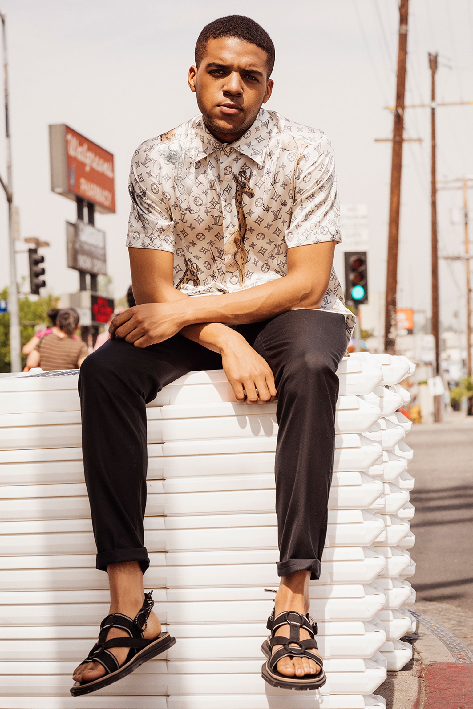 Young man in patterned shirt and black sandals poses on white blocks outdoors in urban setting, captured in a medium shot.
