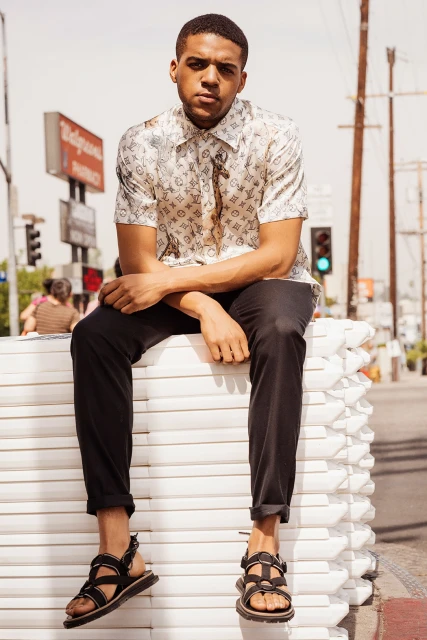 Young man in patterned shirt and black sandals poses on white blocks outdoors in urban setting, captured in a medium shot.