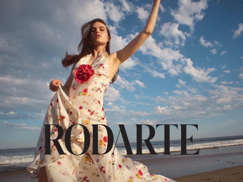 A woman in a floral halter dress stands on a beach under a cloudy blue sky in an outdoor fashion shot by Rodarte.