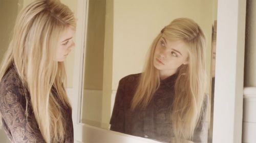Young woman with long blonde hair looking at her reflection in a mirror indoors, wearing a brown patterned shirt.