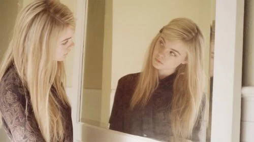 Young woman with long blonde hair looking at her reflection in a mirror indoors, wearing a brown patterned shirt.