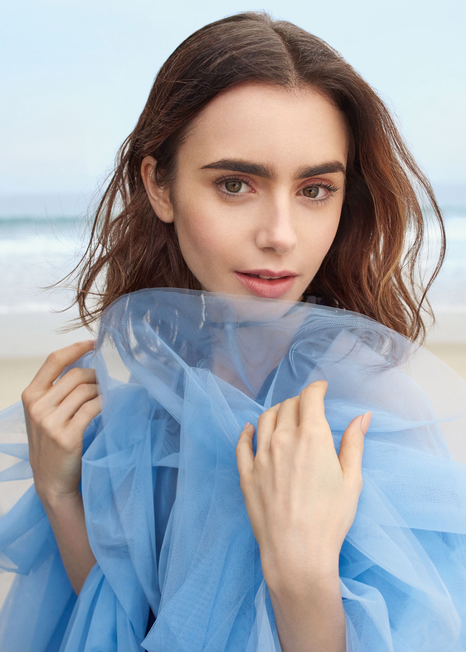 Close-up of an adult woman in a light-blue dress standing on a sandy beach with light skin, brown hair, and neutral facial expression.