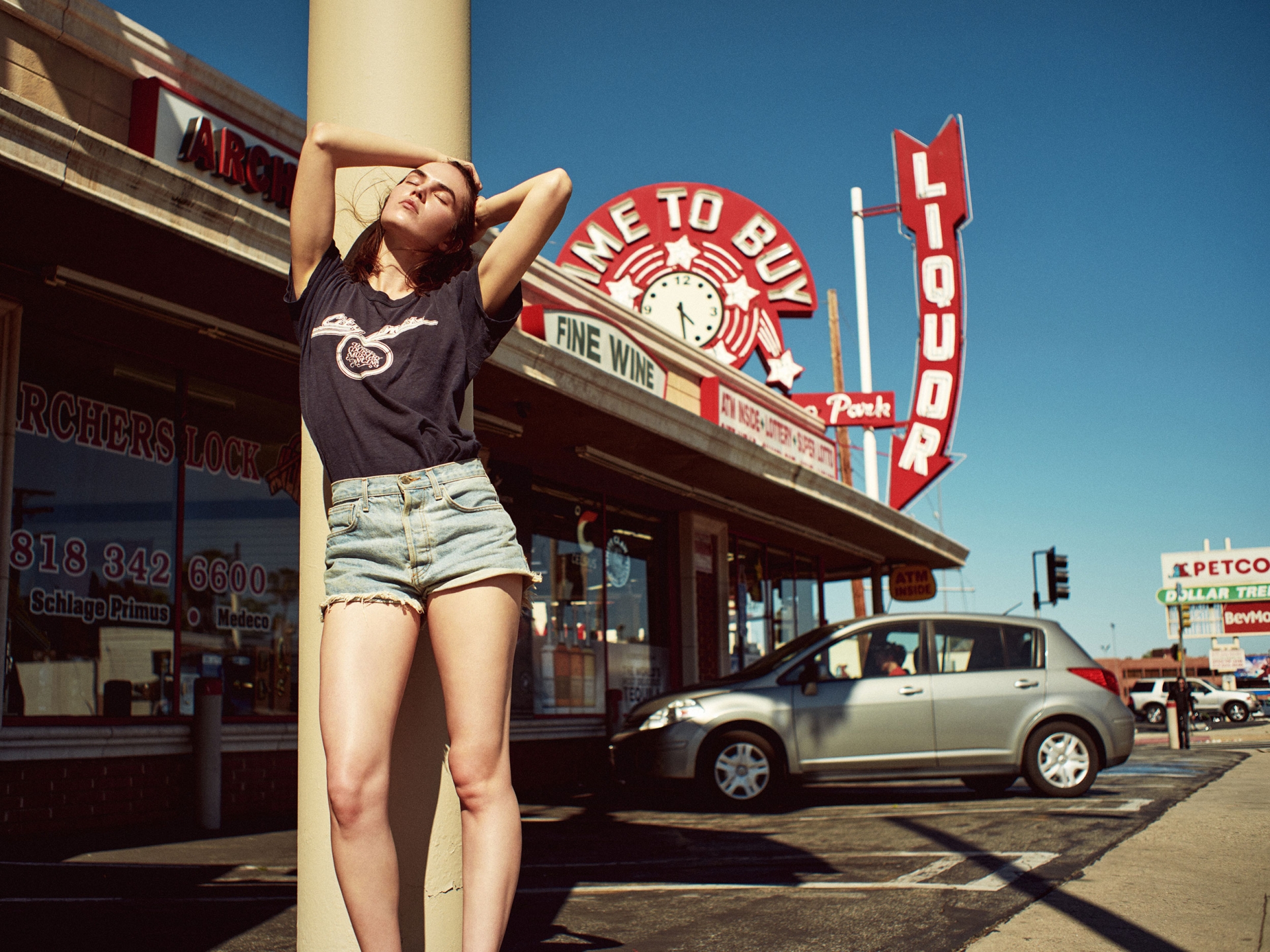 A young adult woman poses outside a liquor store on a sunny day, wearing a dark t-shirt and jean shorts with a relaxed expression.