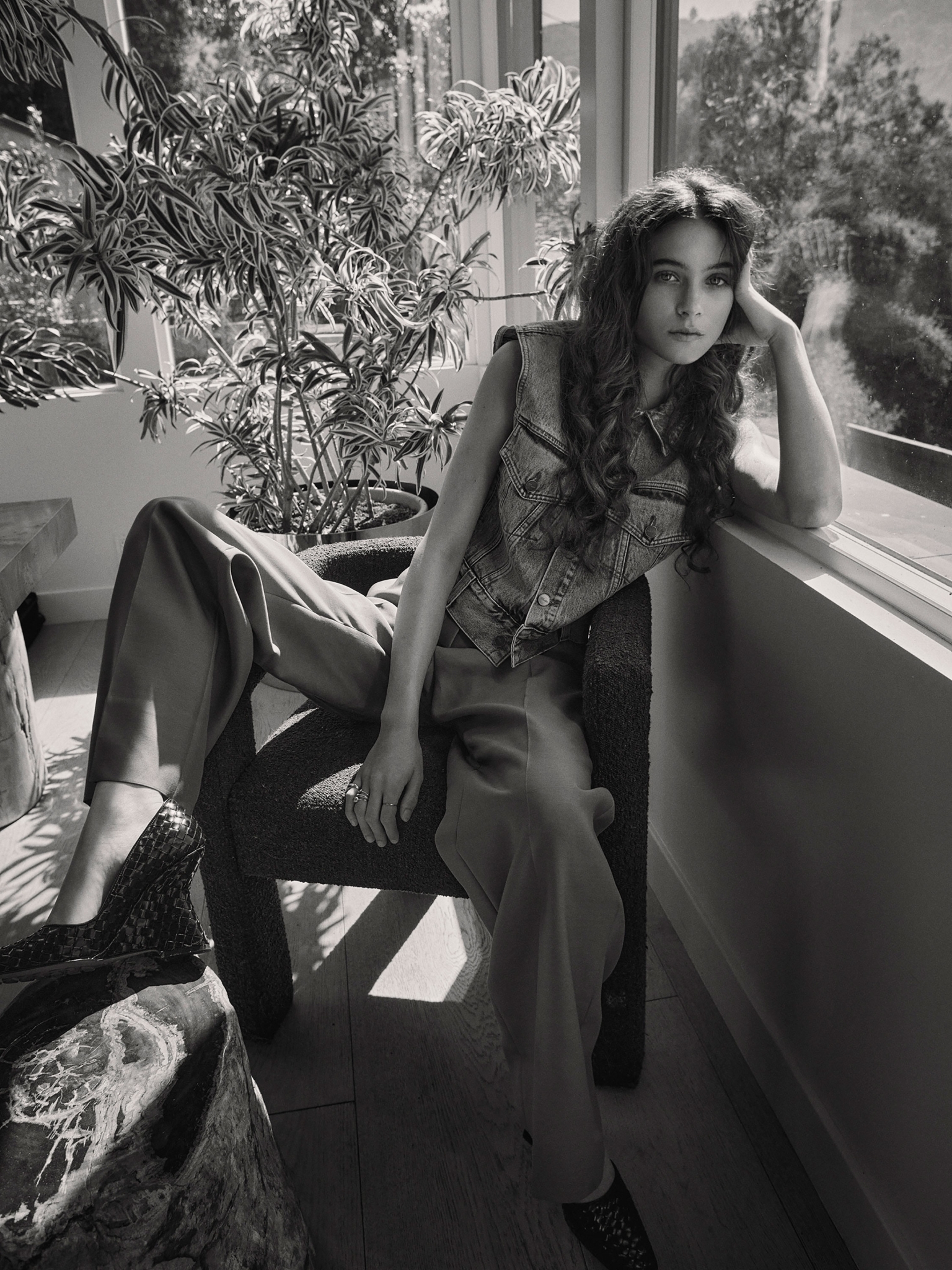 Black and white portrait of a brunette woman in a denim vest and slacks sitting in a chair next to a potted plant by a window.