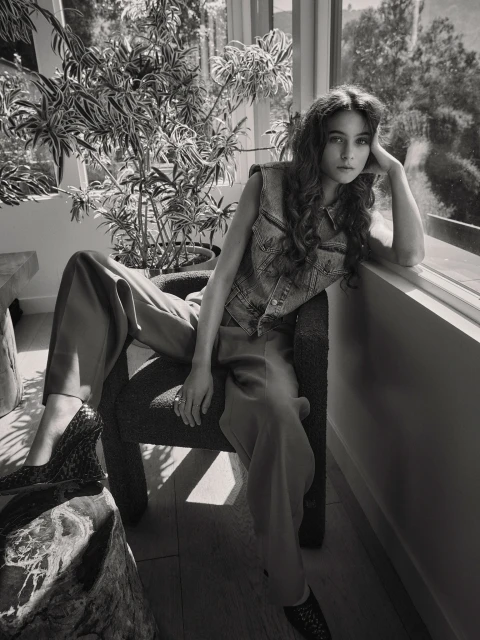 Black and white portrait of a brunette woman in a denim vest and slacks sitting in a chair next to a potted plant by a window.
