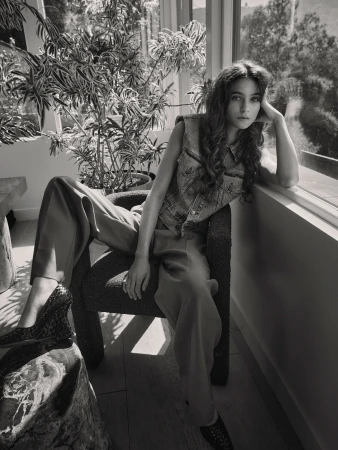 Black and white portrait of a brunette woman in a denim vest and slacks sitting in a chair next to a potted plant by a window.