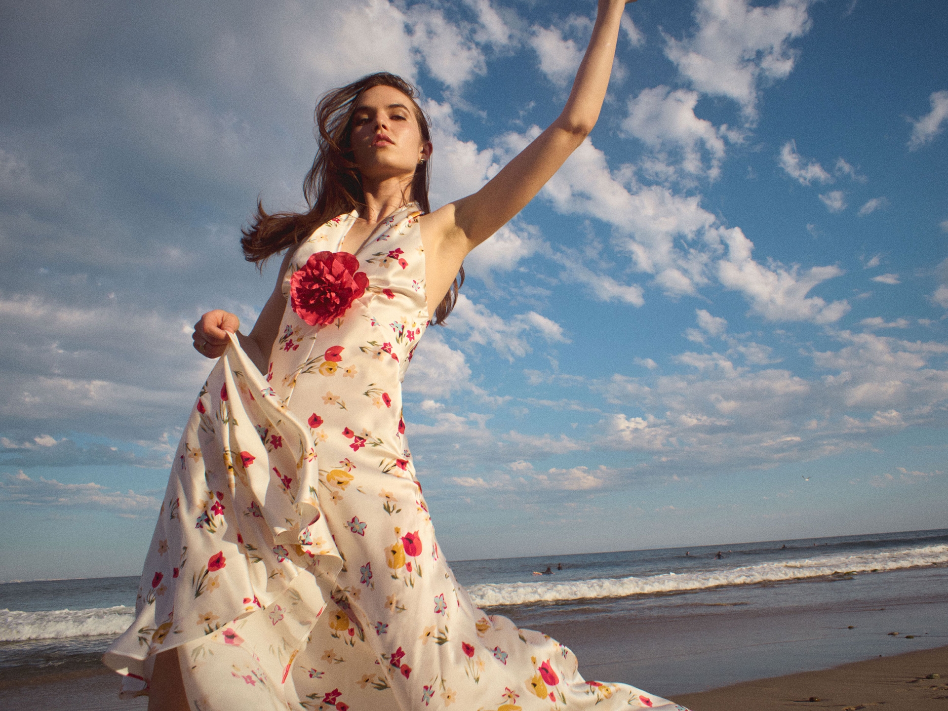 Young woman in floral dress poses on a beach with blue sky and white clouds, her arm raised in a carefree gesture, sunlight.