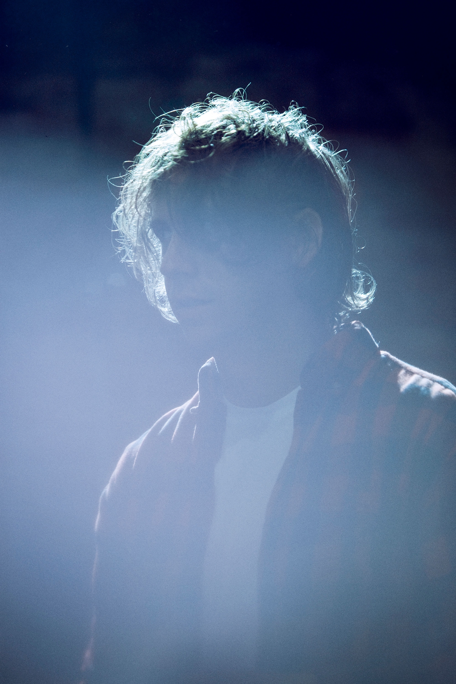 Young person stands backlit in a dark setting wearing a patterned shirt, illuminated by bright white light, with focus on their upper body.