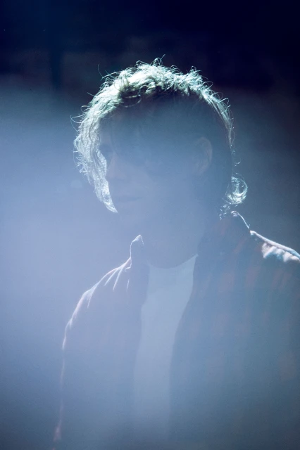Young person stands backlit in a dark setting wearing a patterned shirt, illuminated by bright white light, with focus on their upper body.