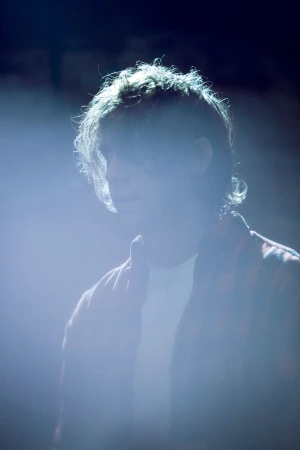 Young person stands backlit in a dark setting wearing a patterned shirt, illuminated by bright white light, with focus on their upper body.