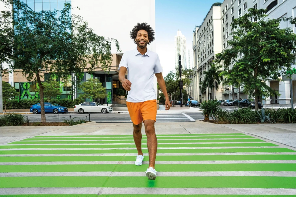 A smiling Black man in casual sportswear walks across a green crosswalk in an urban area with modern buildings and lush trees.