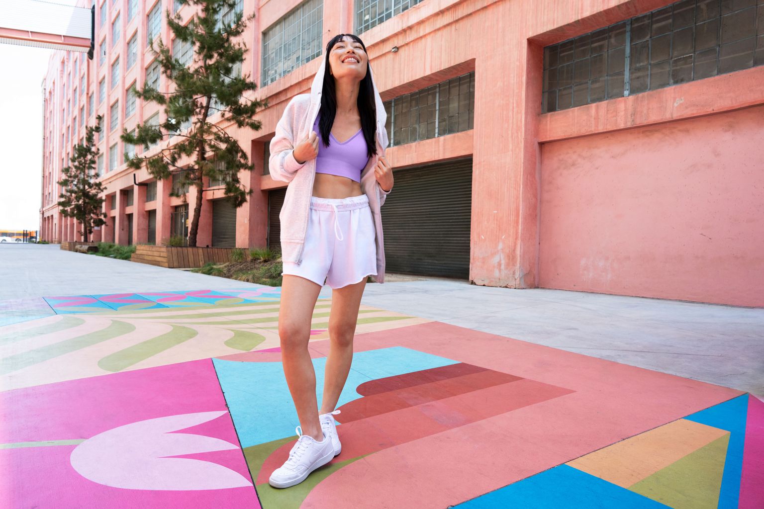 Young Asian woman smiles outdoors in athletic wear, posing in front of a pink industrial building with painted ground art.