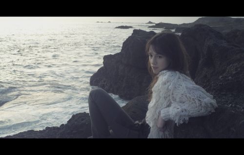 An Asian woman with long hair sits on dark rocks near the ocean. She wears a white sweater and gray pants, looking towards the sea.