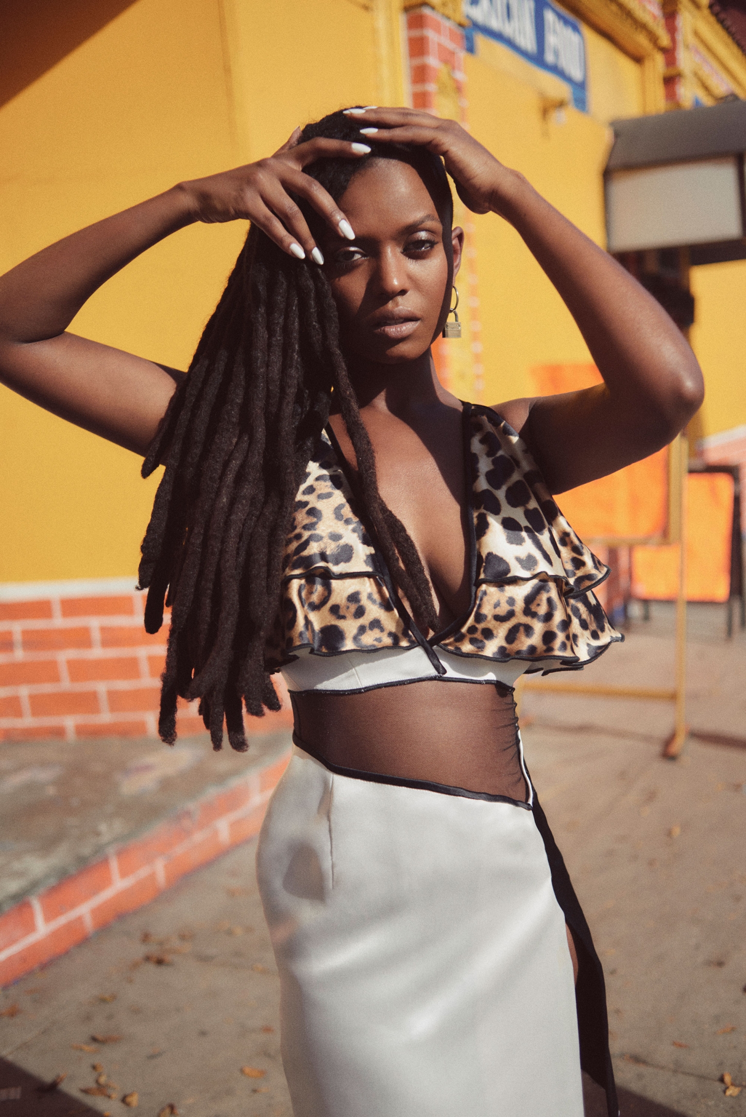 Portrait of a Black woman with dreadlocks wearing an animal-print top and a white skirt, outdoors on a sunny day with a yellow building.