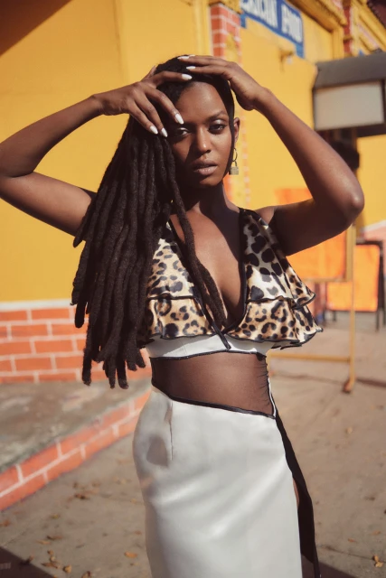 Portrait of a Black woman with dreadlocks wearing an animal-print top and a white skirt, outdoors on a sunny day with a yellow building.