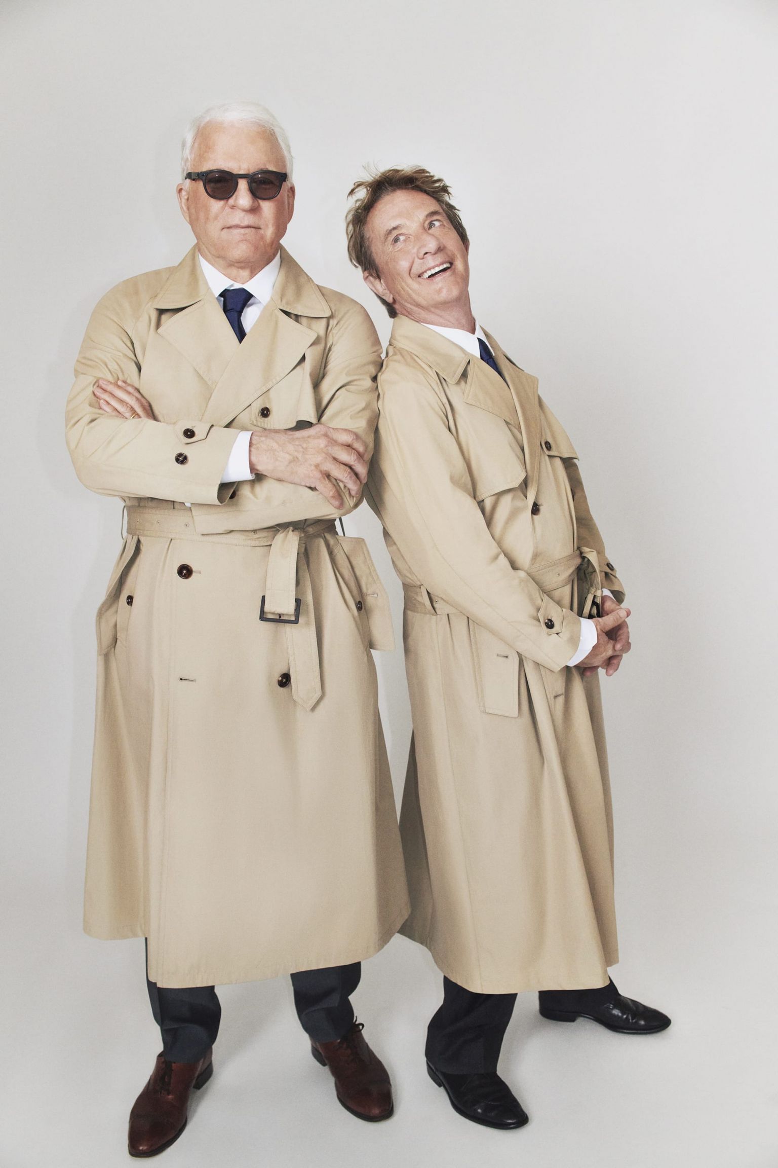 Two adult men, one wearing sunglasses, stand in a light studio setting in tan trench coats, dark trousers and dress shoes in a portrait.