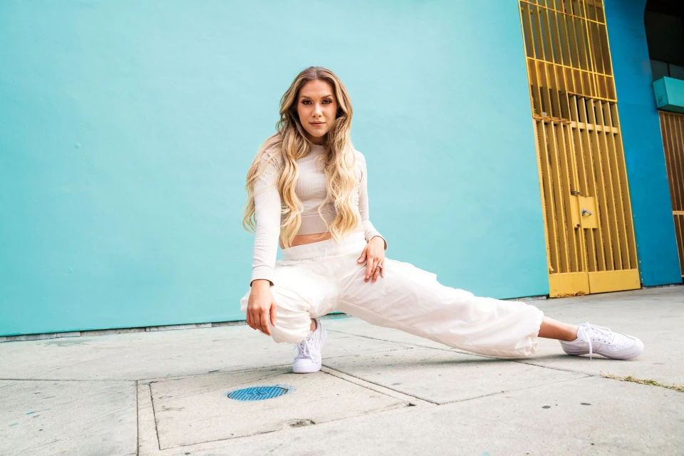 A young adult female with long blonde hair squats in white athletic apparel against a turquoise wall with yellow metal doors.