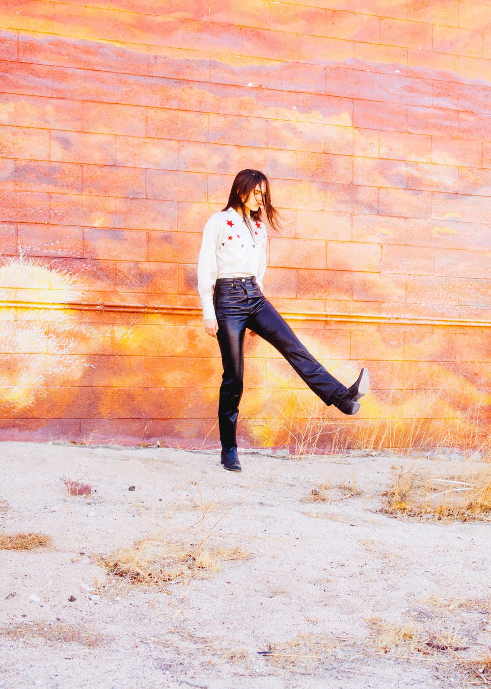 Woman in retro fashion poses against a textured brick wall, wearing a white shirt, black pants, and black boots in an outdoor setting.