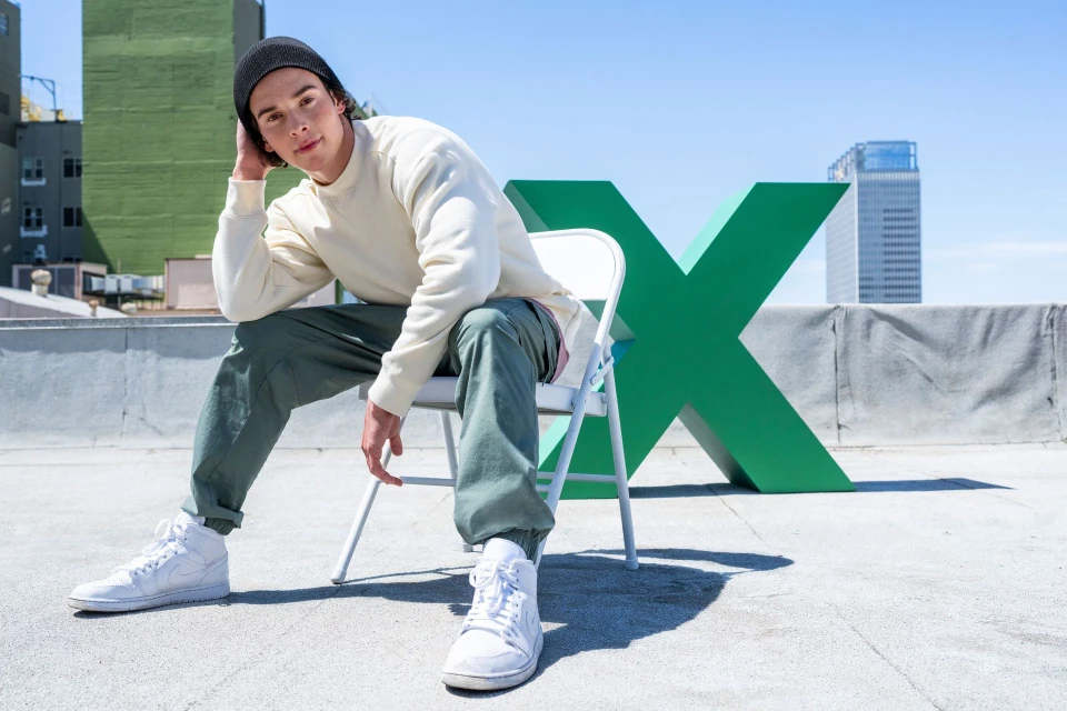 Young man in a beanie sits on a white chair on a rooftop with a large green X, wearing a sweatshirt and sneakers in a sunny urban setting.