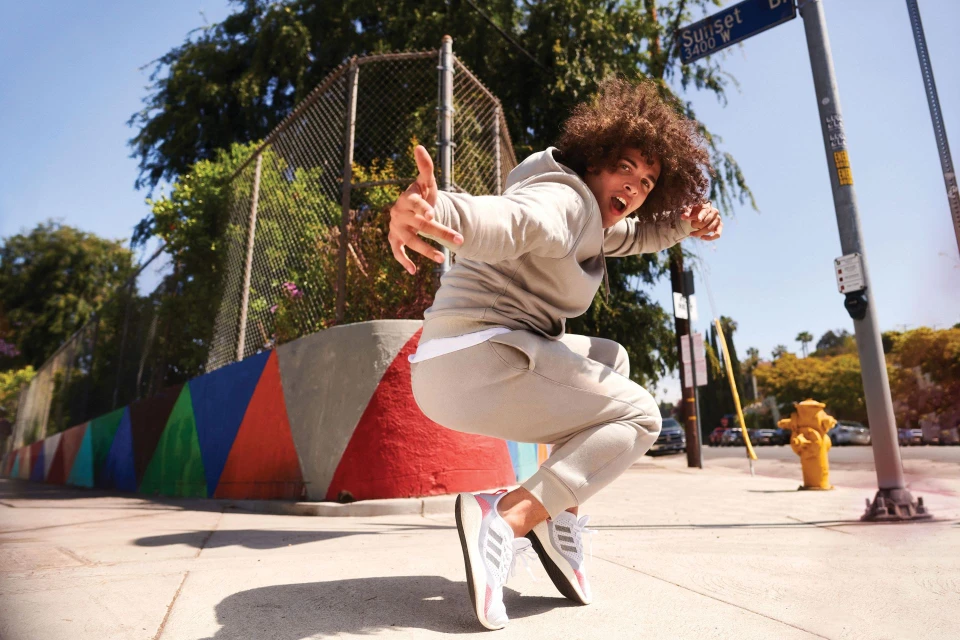 Man in gray sweatsuit leaps on urban street with colorful mural, capturing dynamic energy and athletic style against a sunny sky.