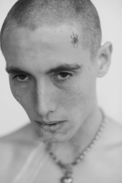 Monochrome portrait of a young adult with a shaved head, sporting a spider tattoo on his forehead, and a beaded necklace with a heart pendant.