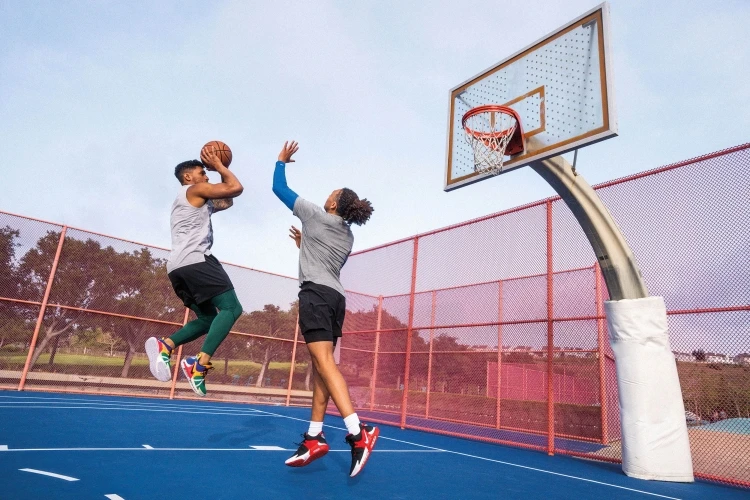 Two adult male basketball players in action on a blue outdoor court with a red fence and a basketball hoop on a sunny day.