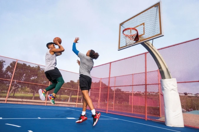 Two adult male basketball players in action on a blue outdoor court with a red fence and a basketball hoop on a sunny day.