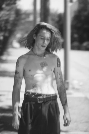 Shirtless young man with long hair and tattoos walking outdoors on a street by Todd Cole