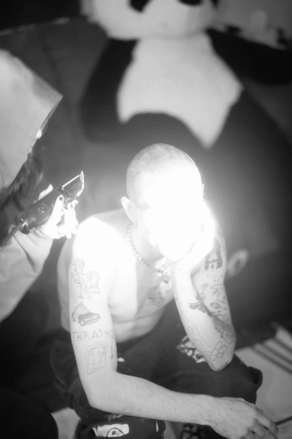Monochrome shot of a shirtless man with tattoos getting a tattoo on his arm, a tattoo artist and a large stuffed panda in the background.