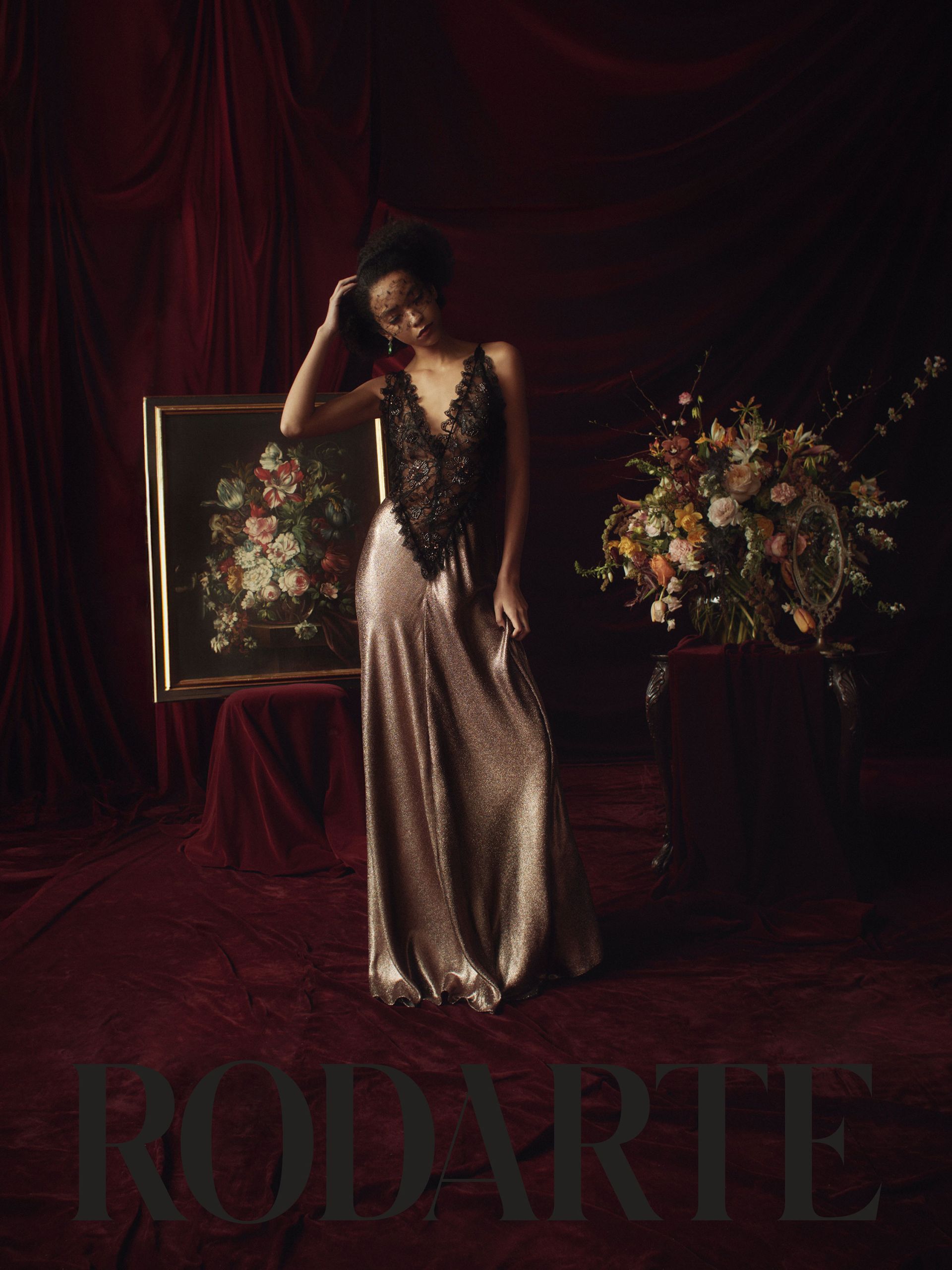 A Black female fashion model poses indoors in a gown with a lace bodice in front of a floral arrangement, vintage painting and velvet curtains.