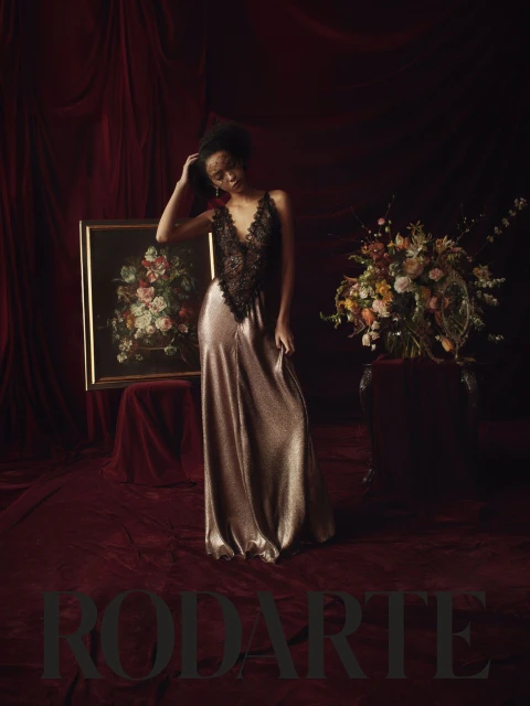 A Black female fashion model poses indoors in a gown with a lace bodice in front of a floral arrangement, vintage painting and velvet curtains.